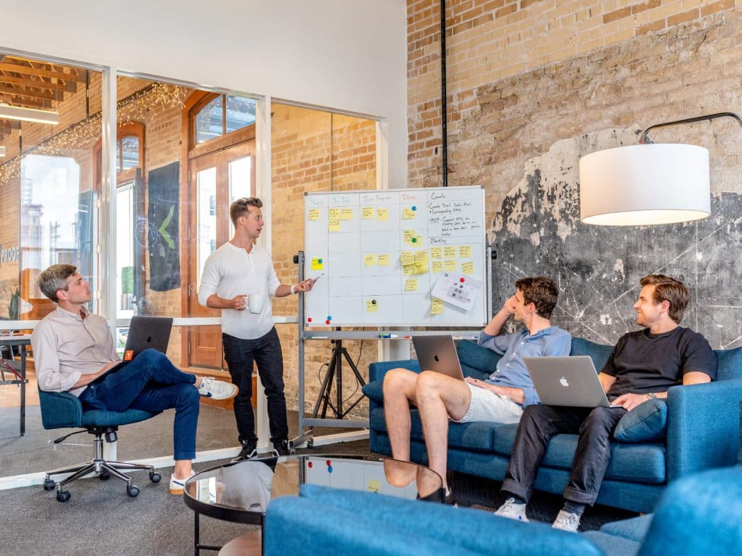 A man stands at a board full of post-its, in front of his co-workers.