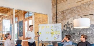 A man stands at a board full of post-its, in front of his co-workers.