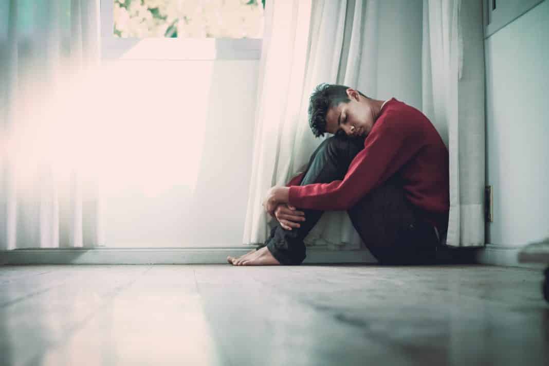 A man sits on a bathroom floor with his knees pulled up to his chest