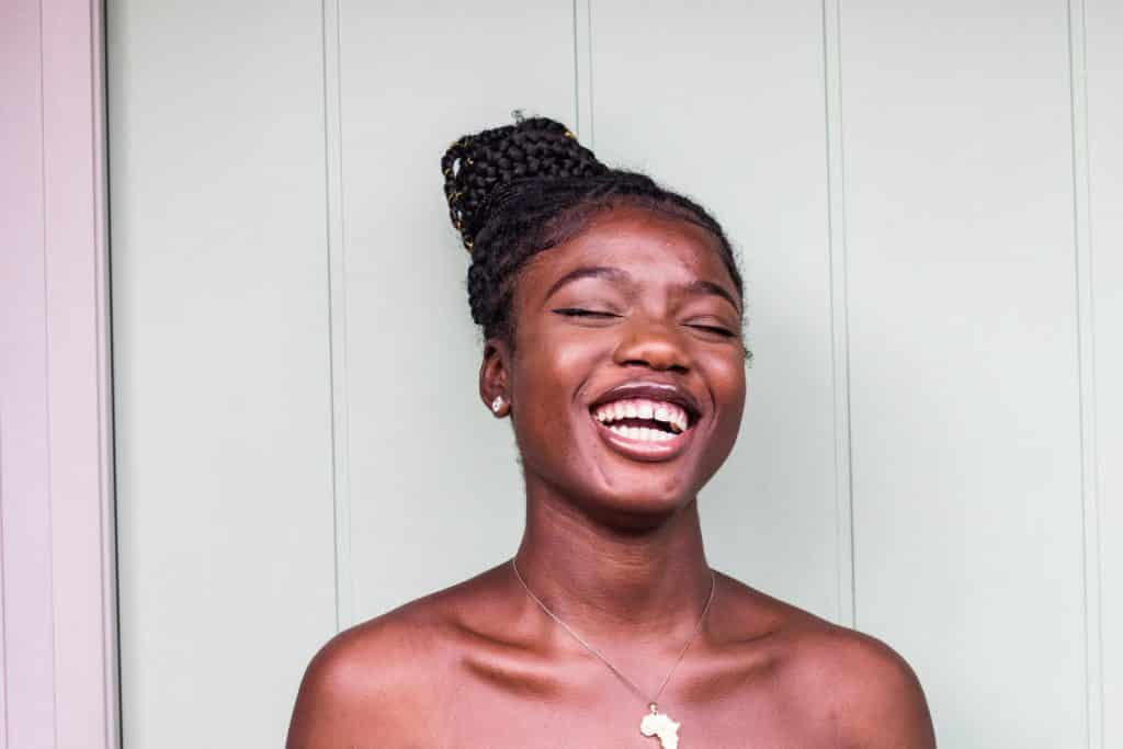 A young woman wearing a necklace shaped like the African continent laughs by a wall
