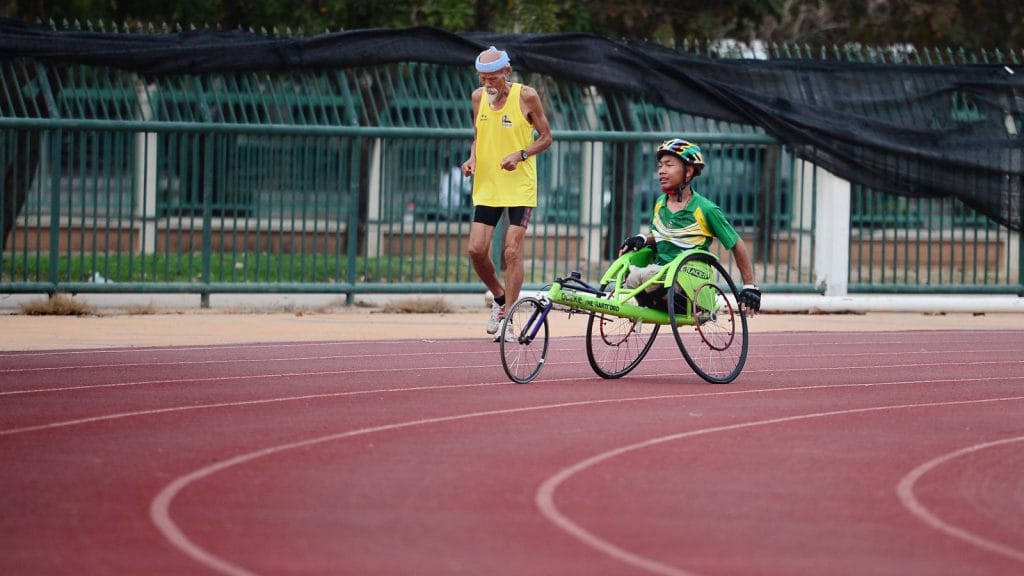 A man in a racing wheelchair races along a running track with an older man running alongside him