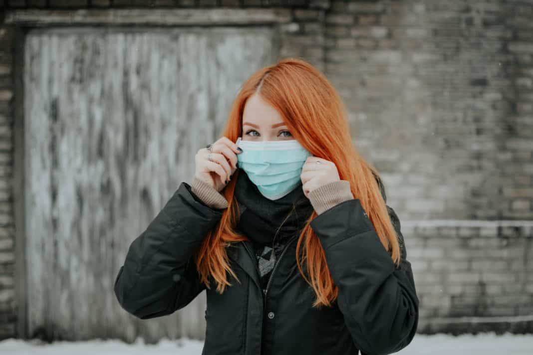A red-haired woman wears a surgical mask over the lower half of her face