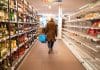 A woman holding a shopping basket walks down a supermarket aisle, with bottles to her left and empty shelves to her right.