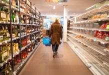 A woman holding a shopping basket walks down a supermarket aisle, with bottles to her left and empty shelves to her right.