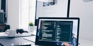 A MacBook with lines of code on its screen on a busy desk